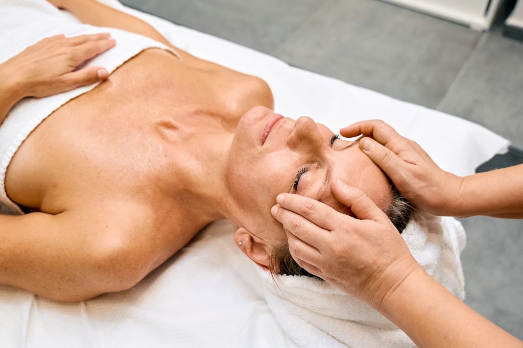 Middle aged woman enjoys hand anti-aging facial treatment massage in spa salon.